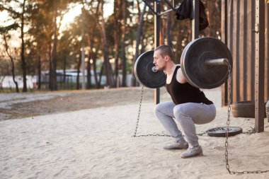Parkın doğasında halterli spor salonu olan adam. Genç beyaz bir sporcu ağır bir ağırlıkla halteri kaldırır ve spor yapar. Sağlıklı yaşam tarzı kavramı. Karantinada spor. Boşluğu kopyala