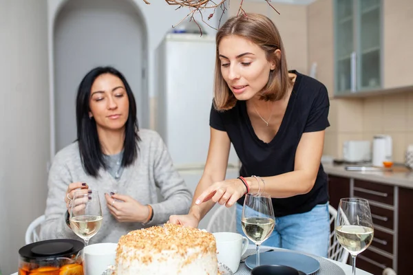 Friends meeting with wine and cake in the modern style kitchen. Women smile and joke. Two  girls drink wine in the home kitchen.