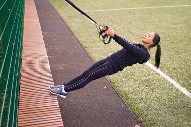 Trx spor sahası kullanan kız sporcu eğitimi. Karışık ırk genç yetişkin kadın spor süspansiyon sistemi. Açık havada esnemek için sağlıklı bir yaşam tarzı. Vücut makineni yap                            