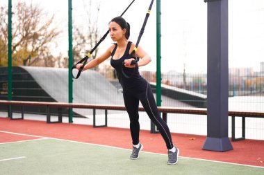 Trx spor sahası kullanan kız sporcu eğitimi. Karışık ırk genç yetişkin kadın spor süspansiyon sistemi. Açık havada esnemek için sağlıklı bir yaşam tarzı. Vücut makineni yap                            
