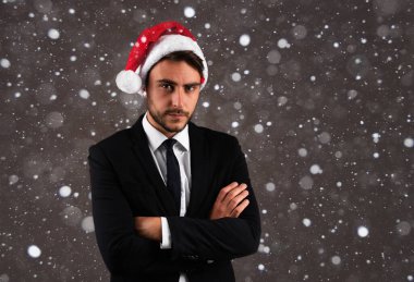 İş elbisesi içindeki yakışıklı beyaz adam ve Noel Baba şapkası stüdyoda gri arka planda duruyor. Ciddi yüzlü, kollarını göğsünde kavuşturmuş, Noel havasında bir iş adamı.