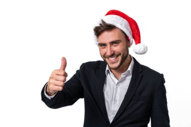 İş elbisesi içindeki yakışıklı beyaz adam ve Noel Baba şapkaları stüdyo gülümsemesinde beyaz arka planda duruyor ve başparmak kaldırıyor. Noel havasında portre yapan iş adamı tatil temalı.