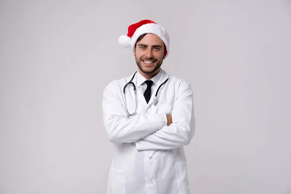 Beyaz üniformalı genç yakışıklı doktor ve Noel Baba şapkası stüdyoda beyaz arka planda duruyor kameraya bakıyor ve diş tebessümü takıyor yeni yıl tıp fakültesi yılbaşı portresi konsepti