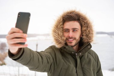 Yakışıklı, sakallı, beyaz, açık havada duran, kürk kapüşonlu, donmuş göl mutlu, çekici Avrupalı adam akıllı telefon fotoğrafçısı Kış Bülteni macerasında selfie çekiyor.
