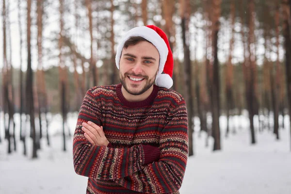 ハンサムな髭を生やした若い白人男性が屋外セーターサンタ帽子冬の森に立っています 雪のクリスマスの森を歩く魅力的なスタイリッシュなヨーロッパの男 シーズン休暇レジャー — ストック写真