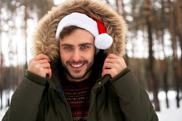 ハンサムな髭を生やした若い白人男性が屋外セーターサンタ帽子冬の森に立っています 雪のクリスマスの森を歩く魅力的なスタイリッシュなヨーロッパの男 シーズン休暇レジャー — ストック写真