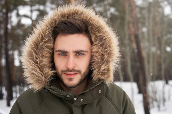 冬の森の中に屋外の毛皮のフードに立ってハンサムな髭を生やした若い白人男性 雪のクリスマスの森を歩く魅力的なスタイリッシュなヨーロッパの男 シーズン休暇レジャー — ストック写真