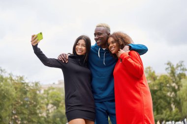 Çok ırklı arkadaşlar dışarıda akıllı telefondan selfie çekiyorlar. Çeşitli gruplar Afro-Amerikan Asyalıların birlikte zaman geçirdiği çok ırklı erkek öğrenci açık havada buluşuyor.