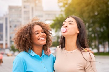 Asyalı arkadaşı sakız şişirirken Afrikalı Amerikalı kadın gülüyor. Açık havada eğlenen etnik çeşitliliğe sahip dostların yakın yüzü. Kız gülüyor ve arkadaşlarıyla sakız çiğniyor. 