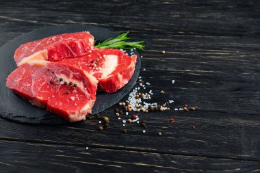 Siyah tahta bir arka planda, taş kesme tahtasına biberiyeli üç parça sulu çiğ biftek. Biber ve tuz çeşnili barbekü ya da ızgara et.