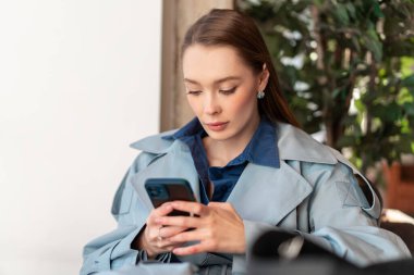 Mavi trençkotlu genç kadın yeşil bitkinin yanında otururken sakin atmosfere ve yumuşak ışığa odaklanmış akıllı telefon kullanıyor.