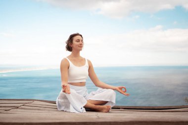 Lotus Poz Padmasana 'yı uygulayan genç bir kadın. Tahta platformda, Mudra' da elleriyle meditasyon yapıyor. Doğada sakinlik, farkındalık ve dengeyi sağlıyor.