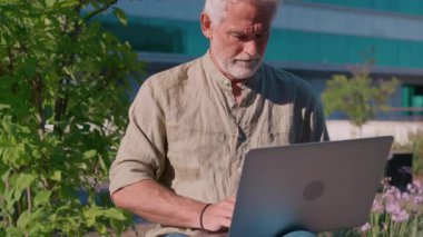 Modern binanın yanında dizüstü bilgisayar kullanan yaşlı bir adam. Günlük tişört giyen bir iş adamı yeşil ortamda uzaktan çalışıyor.