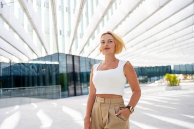 Modern şehir ortamında açık havada duran kısa sarı saçlı, kendine güvenen bir iş kadını. Beyaz bluz ve bej pantolon giyiyor. Güçlü duruş ve olumlu duygu gösteriyor.