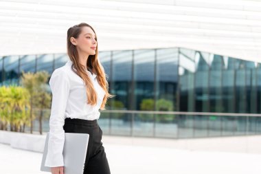 Beyaz gömlekli genç bir iş kadını elinde dizüstü bilgisayarla yürüyor. Uzun kahverengi saçlı, kentteki modern cam binanın önünde kendinden emin görünüyor.