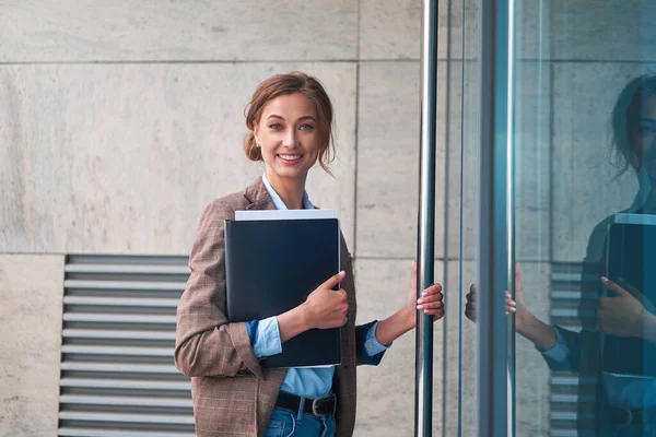 Ofise kimlikleriyle giren, kameraya gülümseyen, kendine güvenen beyaz iş kadını.