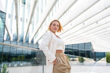 Kısa sarı saçlı, spor ceketli ve pantolonu parmaklıklara dayanmış genç bir iş kadını. Gündüz vakti modern ofis binasının dışındaki kameraya bakıyor.