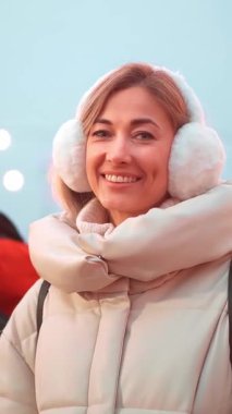 Cheerful woman dressed in a cozy winter coat and earmuffs smiles amidst holiday decorations and glowing lights during a crisp winter evening celebration.