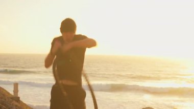 Determined sporty man practicing air boxing with battle ropes on platform by sea against sky during sunset. Young fit male doing crossfit training in nature