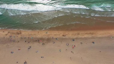 Aerial drone top down view of beachgoers relaxing on sand with sea waves crashing along the coastline on sunny summer day