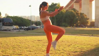 Woman performing high knee exercise in park near bridge during sunset. Wearing sports bra and orange leggings, she is staying fit in park during workout session