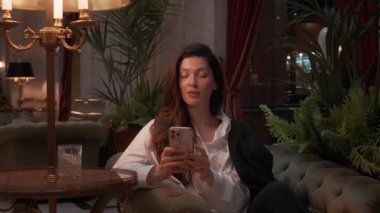 Business woman checking smartphone while relaxing on sofa in luxurious hotel lobby with warm lighting and elegant interior decor in the background