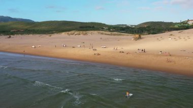 Aerial drone view of surfers with waves breaking on shore at Guincho Beach, Portugal. Rolling hills and sandy beach provide scenic backdrop for active surfing scene