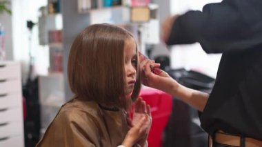 Cute girl getting haircut by hairdresser in salon. Female stylist creates hairstyle using comb. The stylist takes care of the child hair at barbershop