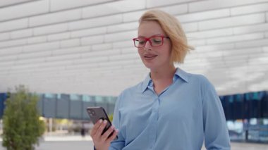 Young businesswoman with short blond hair and red glasses watching video on smartphone while standing near modern office building