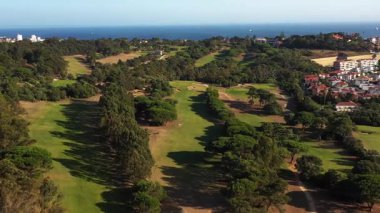 Aerial drone captures stunning view of golf course bordered by lush green trees on sunny day, creating serene and scenic landscape with natural beauty