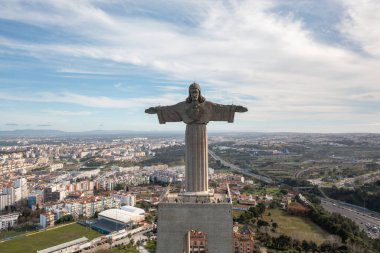 Aerial drone shot of iconic Christ the King statue in Almada, Portugal with arms outstretched overseeing the cityscape under bright sky