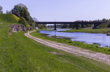 dolambaçlı bir nehir bir köprüden ile; Nehir kenarı boyunca boş, çakıllı kırsal yol yoktur; Eflatun çiçekli çalı bir tepenin üzerinde bir o kaldı