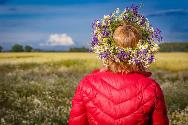 summer solstice, girls\' head wreath made of field meadow flowers; girl in red jacket with floral wreath in head on blurred countryside background