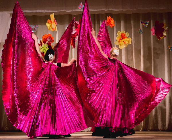 восточный танец на сцене в ярких цветах в фиолетовых тонах с широкими крыльями для болельщиков; вид со спины на танцоров
