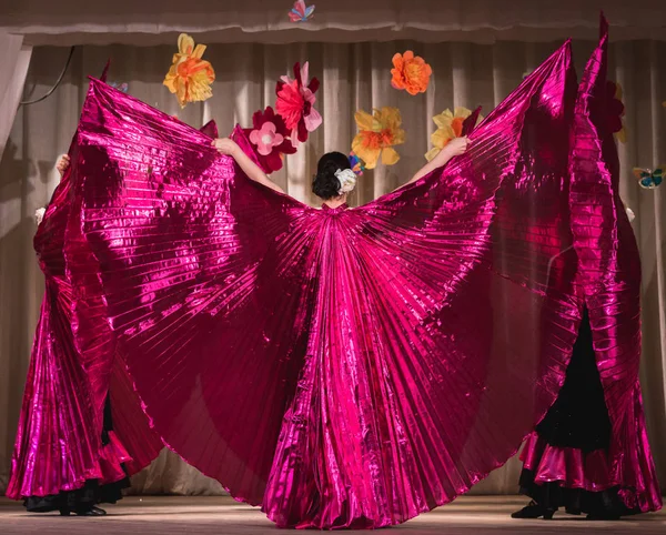 oriental dance on stage in bright colors in purple tones with wide wings for fans; view from the back to the dancers