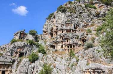 Antik, tarihi bir yer, dağlarda inşa edilmiş eski mezarlar; ön planda onlara kabartmalar ile kayalar ve arka büyük bir dağ