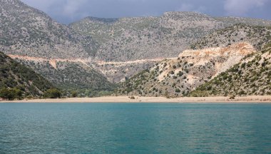 büyük, gri bir dağın eteklerinde uzak bir kumlu plajda bir yat görünümü; deniz ve dağ manzarası