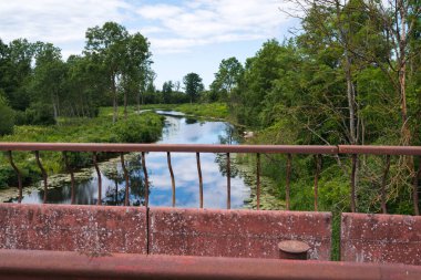 Sudaki bulutların yansımasına sahip küçük, dar bir nehir; kıyılarda ağaçlar ve çalılar var.