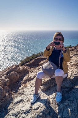 Yolculuk boyunca okyanus kıyısında bir kadın oturuyor, arka planda kayalık bir kıyı, su ve gökyüzü.
