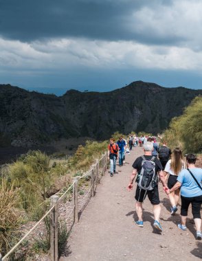 Napoli, İtalya 04.28.2025: Vezüv Dağı 'nın kraterini görmeye giden turistler. Hem yukarı hem de aşağı uzun ve zor bir tırmanış. Arka planda koyu mavi fırtınalı gökyüzü.