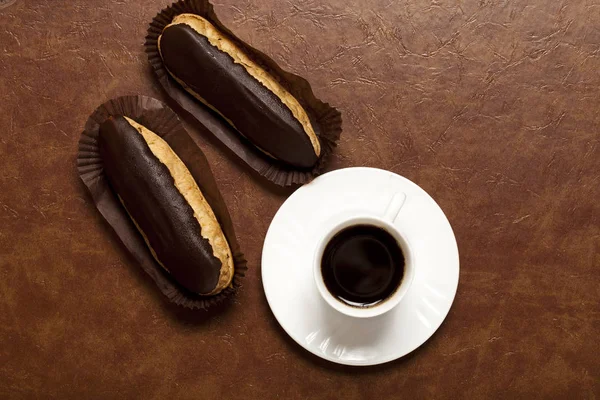 kahve, çikolata Eclair, beyaz bir fincan kahve, beyaz tabak, kahverengi bir masada, bir kağıt standı üzerinde iki eclairs.