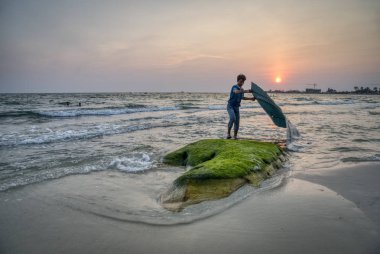 Sihanoukville ,Kamboçya. 26 Şubat 2019: Krong Vaaz Sihanouk plajında güneş batarken saman şiltesini kayanın üzerinde yıkayan genç çocuk.