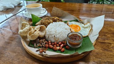 Bir tabak muz yaprağı nasi lemak pilavı, kızarmış yumurta, bitkisel veya kurutulmuş körili tavuk eti, ançüez, baharatlı sos ve balık krakeri.. 
