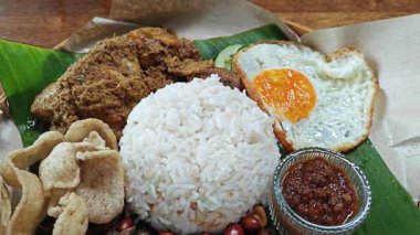 Bir tabak muz yaprağı nasi lemak pilavı, kızarmış yumurta, bitkisel veya kurutulmuş körili tavuk eti, ançüez, baharatlı sos ve balık krakeri.. 