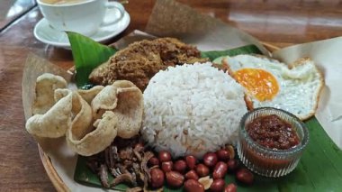 Bir tabak muz yaprağı nasi lemak pilavı, kızarmış yumurta, bitkisel veya kurutulmuş körili tavuk eti, ançüez, baharatlı sos ve balık krakeri.. 