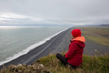 Kırmızı ceketli bir kız, deniz kıyısındaki bir kayalıkta oturur ve ufukta kurşunlu bir gökyüzüyle uzanan siyah lav kumu ile oturur..