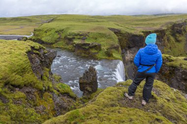 Mavi ceketli bir kız Fagrifoss Şelalesi'nin kenarındaki bir uçurumun kenarında duruyor.