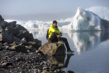 sarı ceketli bir adam, bir turist buzlu lagünün kıyısında bir taş üzerinde oturur