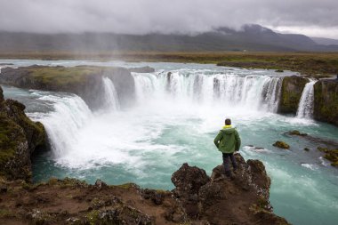 Bir erkek turist bir şelale üzerinde bir uçurumun üzerinde duruyor ve güzelliği hayran. İzlanda'da çekilen fotoğraf.