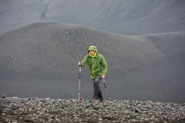A male tourist in a green jacket with trekking poles climbs the side of a sleeping volcano